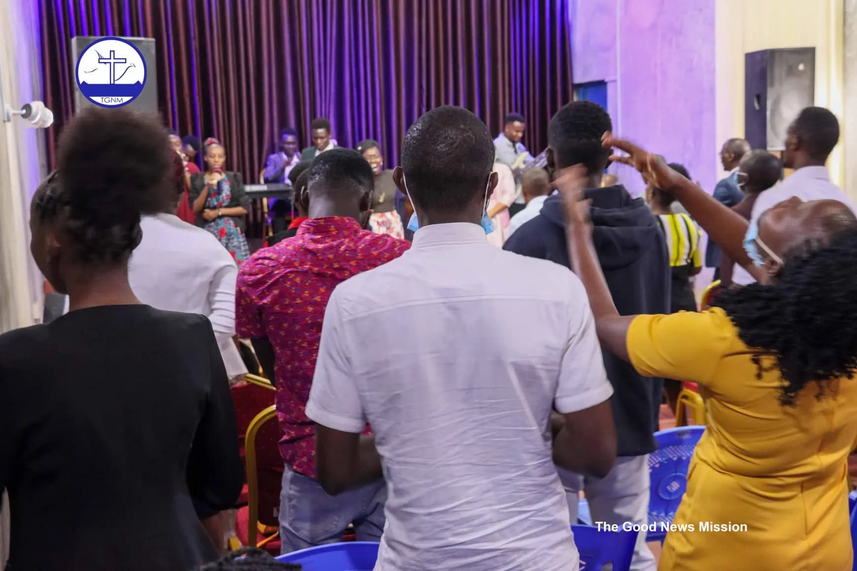 The congregation of The Good News Mission standing in worship with hands raised, a worship leader singing at the front